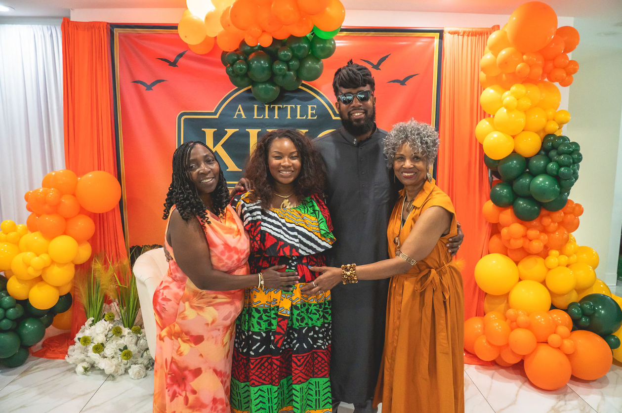 Four family members smiling together at a celebration surrounded by orange, yellow, and green balloons