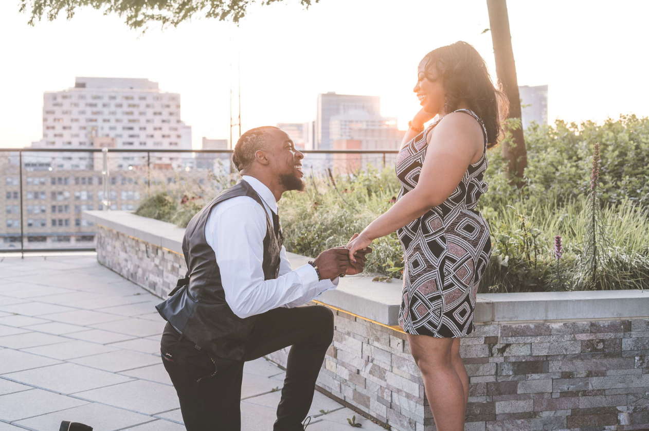 A rooftop proposal with one partner kneeling and holding the other’s hand in the evening light
